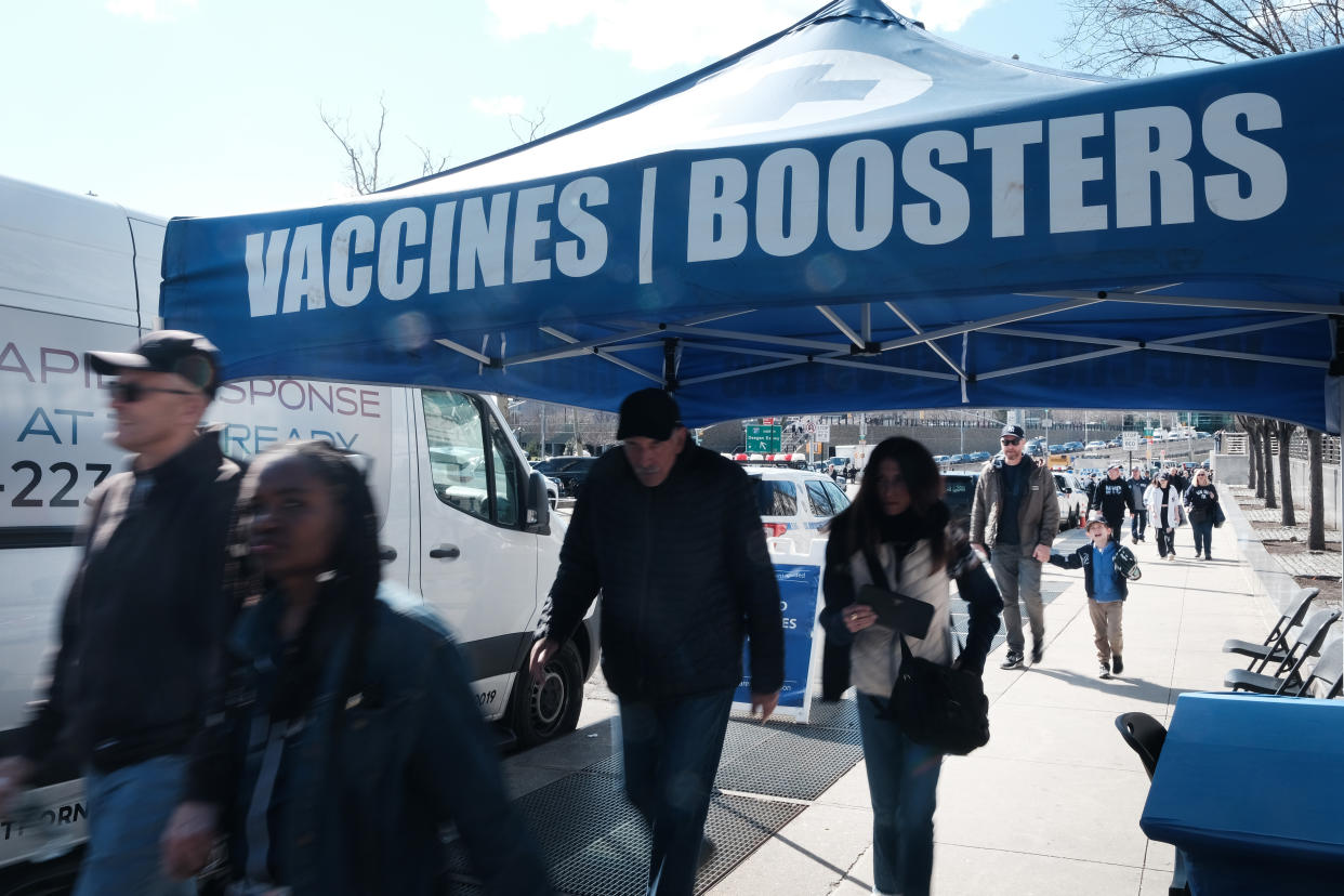 Sidewalk pedestrians pass under an awning that reads: Vaccines/Boosters.