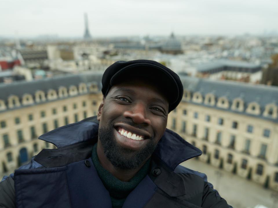 omar sy in lupin, wearing a coat and cap and smiling widely. paris is in the background