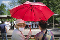 Two women, one wearing a face mask to combat the spread of the coronavirus, shield themselves from the sun under an umbrella while watching a folk group perform a summer solstice dance at the DImitrie Gusti Village Museum in Bucharest, Romania, Saturday, June 27, 2020. (AP Photo/Vadim Ghirda)