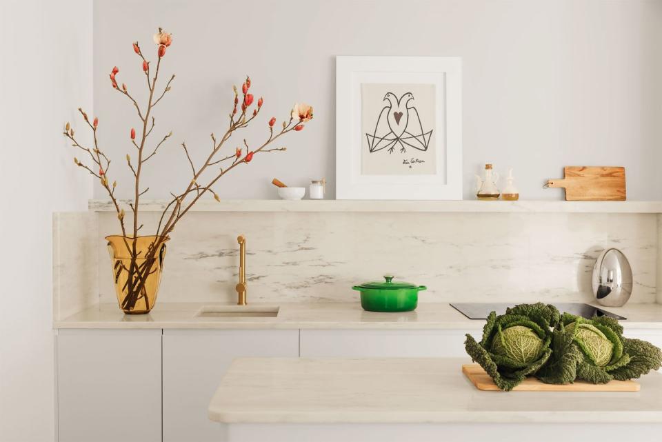 in the kitchen, an island and counter, with a built in sink, are topped with portuguese rosa marble, on the counter is a vase with tall salmon colored buds and a green saucepot, on a shelf is a drawing of two birds