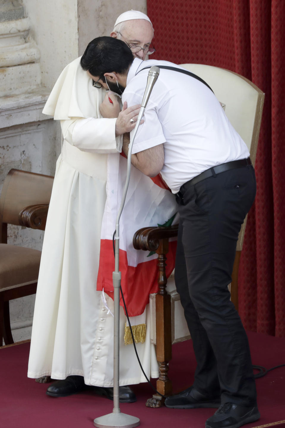 Pope Francis is hugged by Lebanese priest Georges Breidi as they hold a Lebanese flag in remembrance of last month's explosion in Beirut, during the pontiff's general audience, the first with faithful since February when the coronavirus outbreak broke out, at the San Damaso courtyard, at the Vatican, Wednesday, Sept. 2, 2020. (AP Photo/Andrew Medichini)