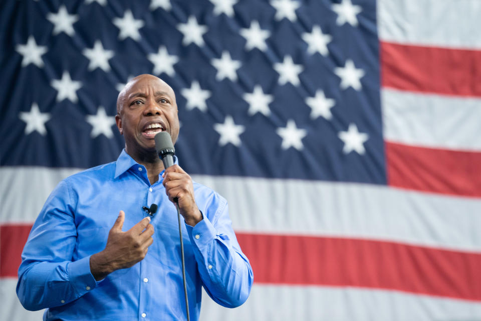 Sen. Tim Scott speaks into a microphone in front of an American flag.