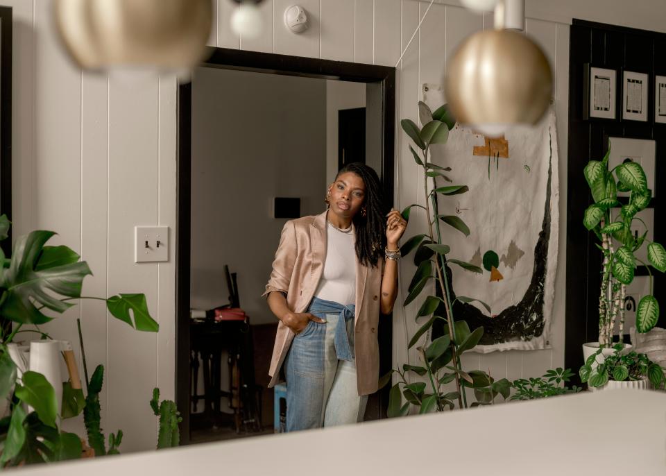 Interior designer Carmeon Hamilton leans on a counter in her home. She's wearing a blush pink blazer over a white shirt with blue jeans and white loafers. Her home is beautifully decorated with plants, paintings, and geometric furniture and fixtures.