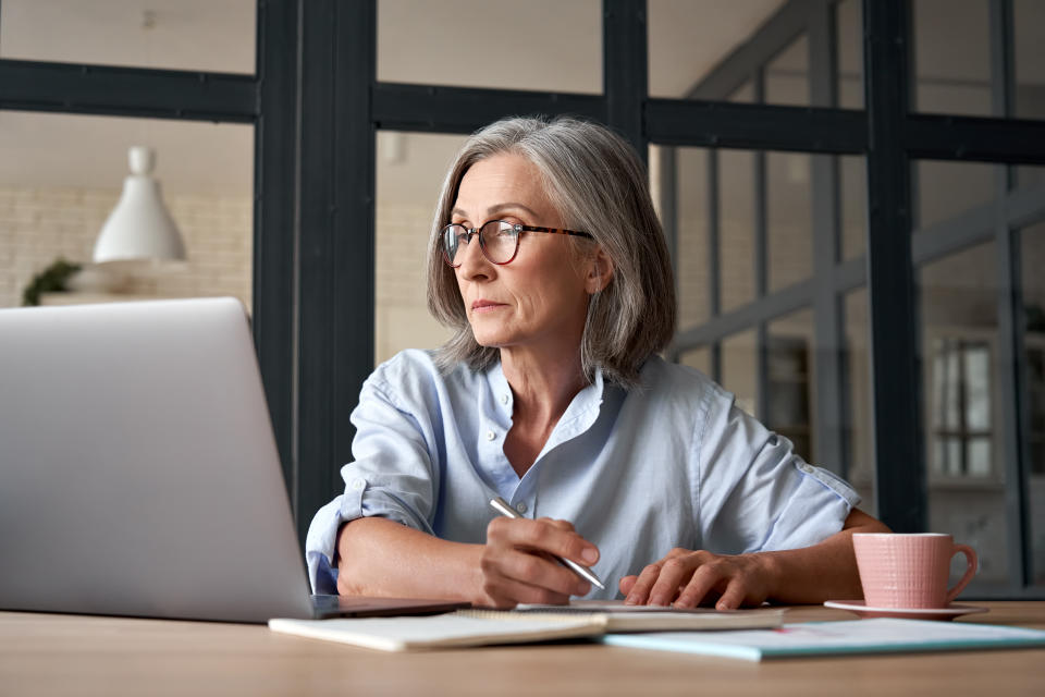 Serious mature older adult woman watching training webinar on laptop working from home or in office. 60s middle aged businesswoman taking notes while using computer technology sitting at table.