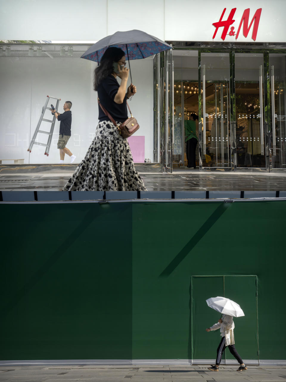 In this combination image, a worker carries a ladder as he works on a window display at an outlet of Sweden-based clothing company H&M on May 16, 2023, top, and a woman carrying an umbrella walks past the now-closed store at a shopping mall in Beijing Wednesday, June 21, 2023. Foreign companies are shifting investments and their Asian headquarters out of China as confidence plunges following the expansion of an anti-spying law and other challenges, the European Union Chamber of Commerce in China said Wednesday. (AP Photo/Mark Schiefelbein)