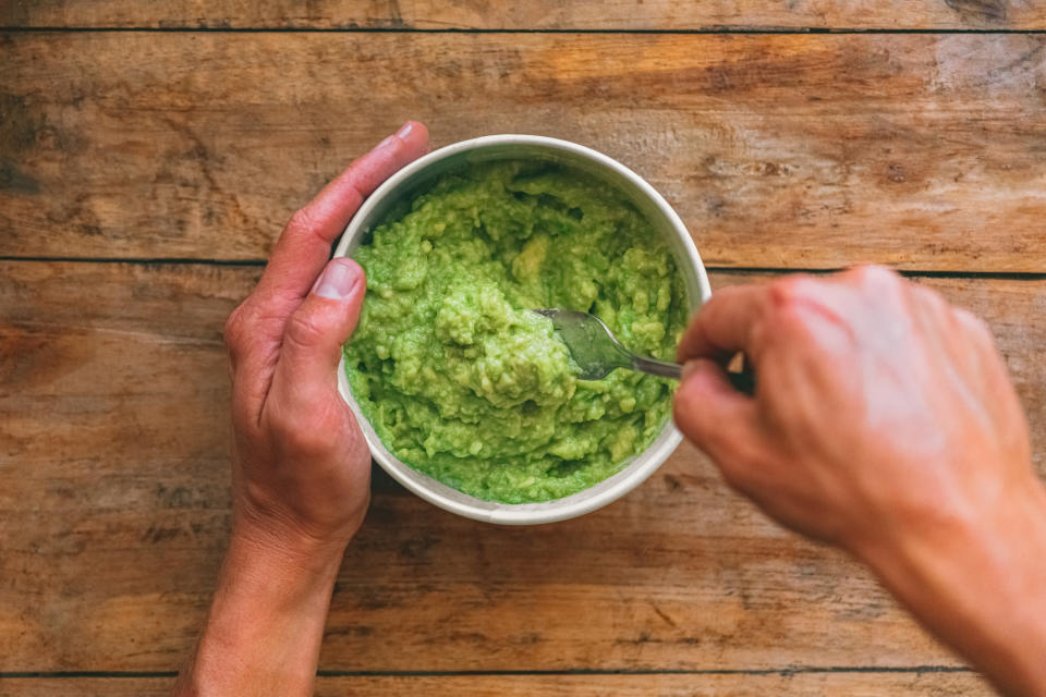 Hands holding a bowl of mashed green avocado with a spoon on a wooden table