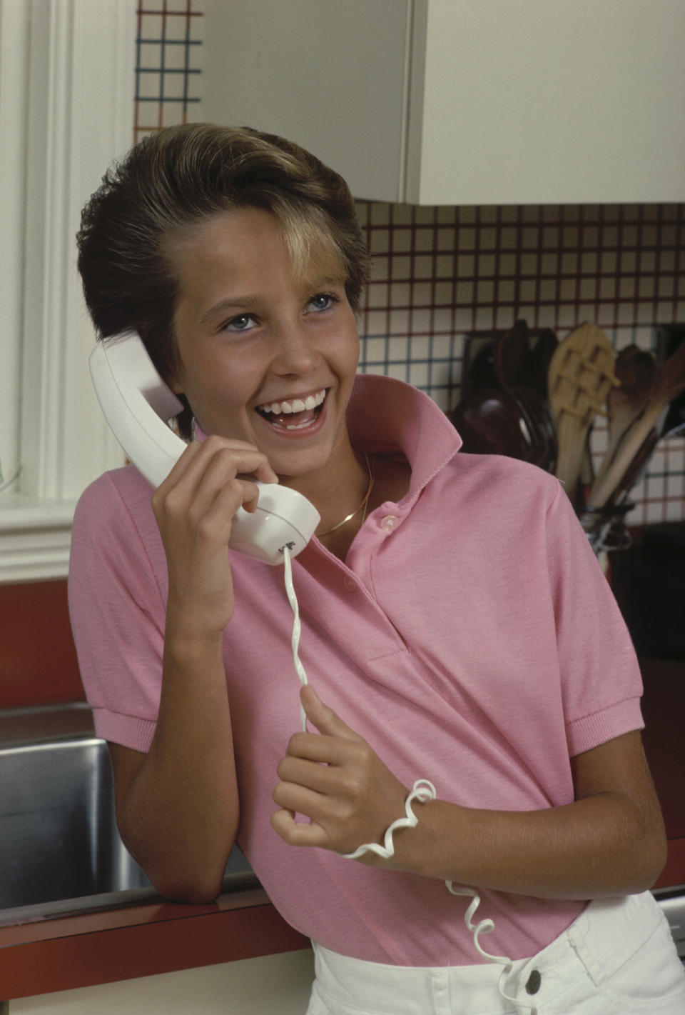 Smiling person holding a telephone receiver, wearing a pink top and white pants, in a kitchen setting