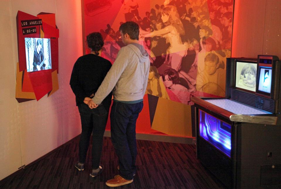 This March 26, 2012 photo shows visitors watching a display about the 1960s 'Go-Go' scene in Los Angeles, next to a vintage Rock-Ola jukebox converted to digital use, at the exhibit, "Trouble In Paradise: Music and Los Angeles, 1945-1975," at the Grammy Museum in Los Angeles. The museum website says the exhibit focuses on the "tensions between alluring myths of Southern California paradise and the realities of social struggle that characterized the years following WWII." (AP Photo/Reed Saxon)