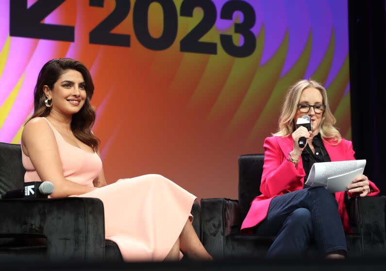 A seated woman in a dress smiles as a seated woman in a pink jacket reads into a mic while holding a sheaf of papers