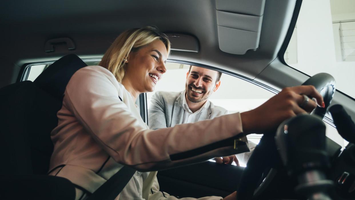 Woman buying a car in dealership sitting in her new auto, the salesman talking to her in the background.