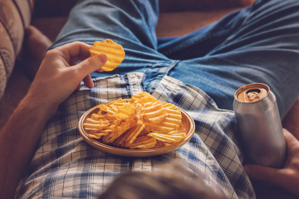 After work a guy wearing shirt and jeans lying on sofa, drinking a cold beer, eating crisps and watching sport tv channel. Man's resting time at home concept.