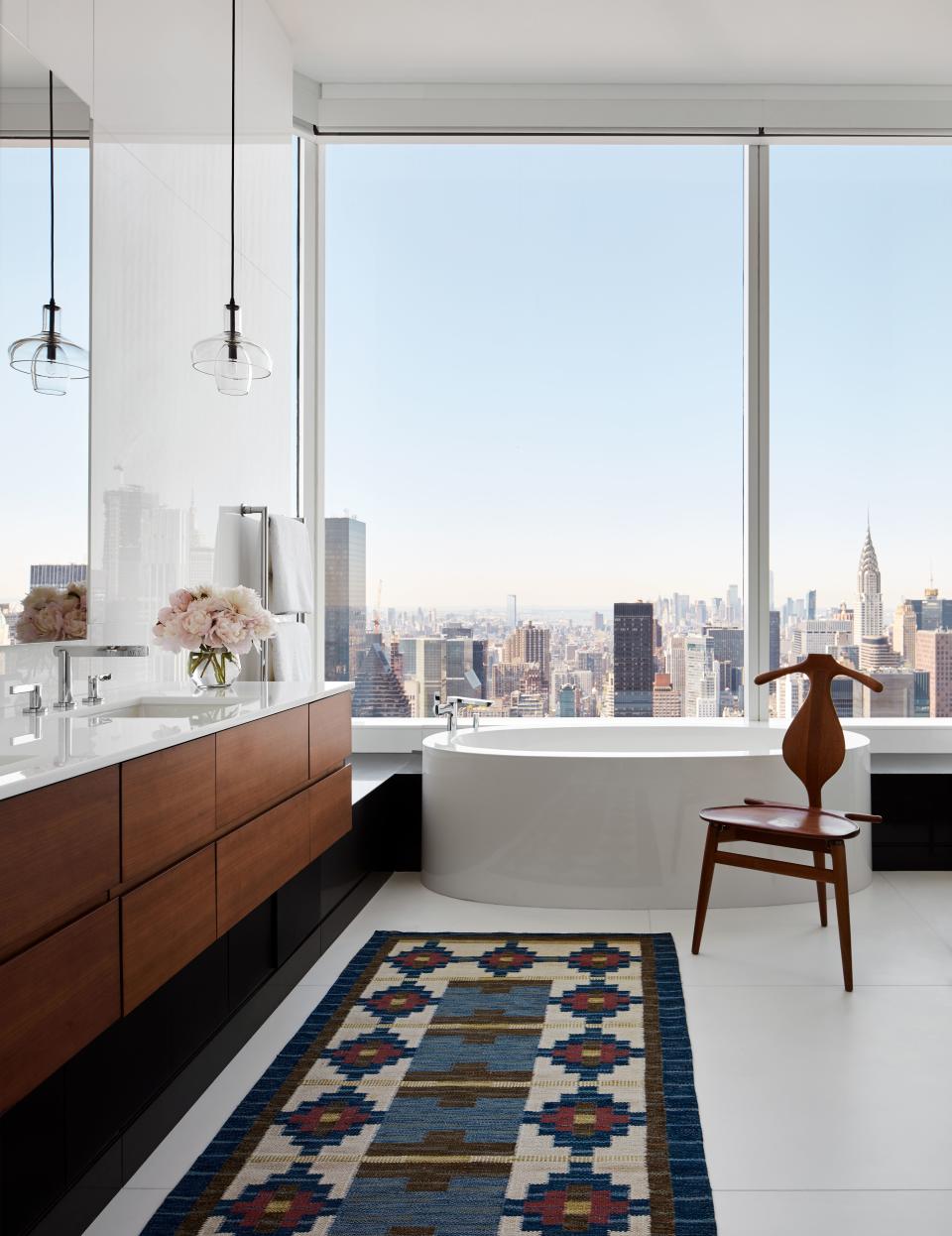 The serenity of the master suite carries over to the stunning bathroom, boasting a sculptural tub designed by Daniel Romualdez that offers bathers a bird’s-eye view of Midtown Manhattan. Polished white nanoglass tiles the walls and floors, warmed by a vintage Swedish flat weave rug. Wooden accents come from the vanity and a vintage carved teak valet chair by Hans Wegner. The pendant lights are by Alison Berger.
