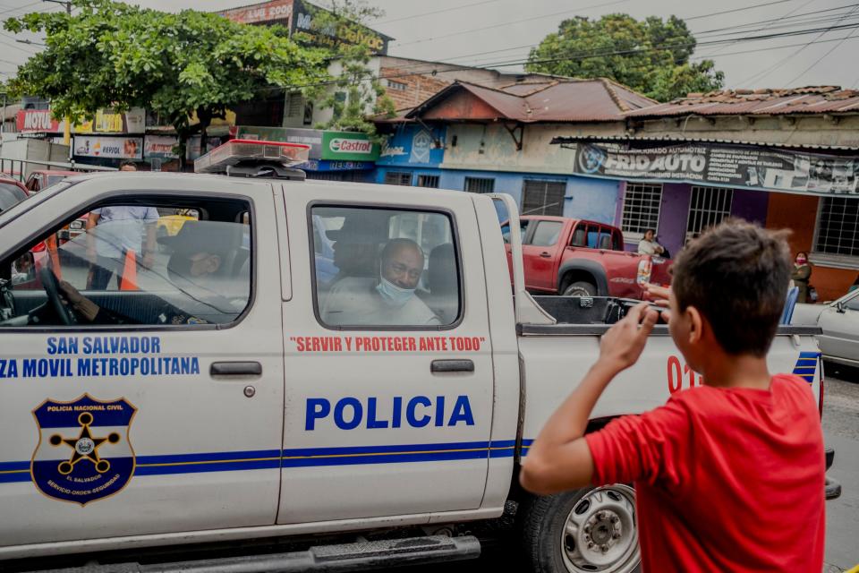 A boy looks on as a man sits in the back of a police car.