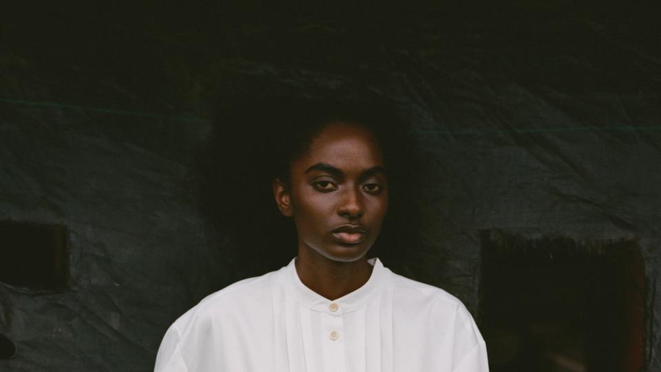 a model in a white shirt dress with a wooden carved human heart worn around her waist poses in front of a black background