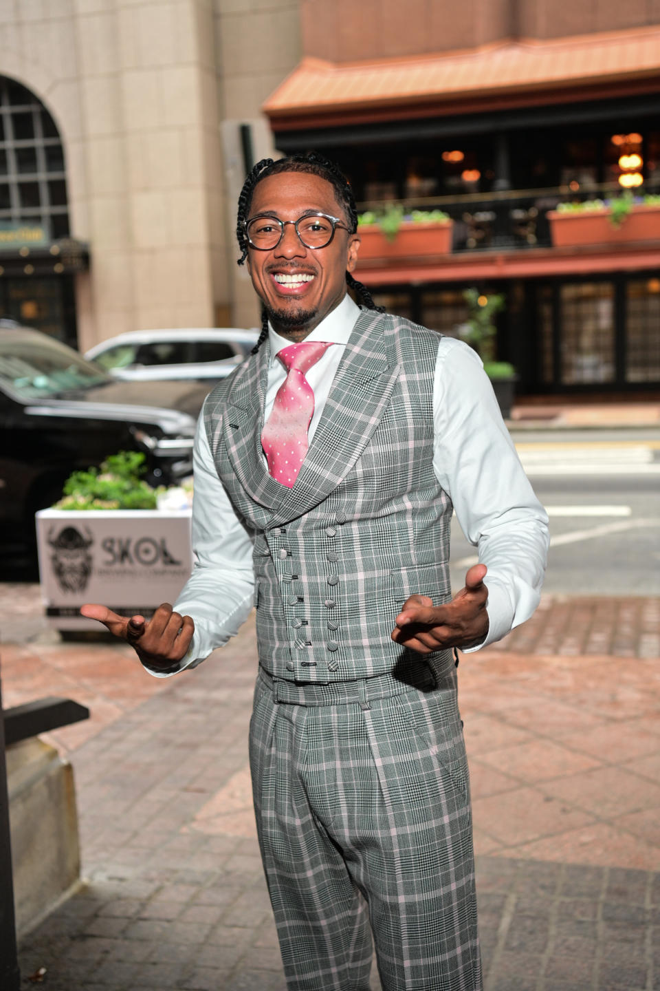 Nick Cannon in a grey checked suit with a pink tie, smiles and gestures with his hands on a sidewalk in the city
