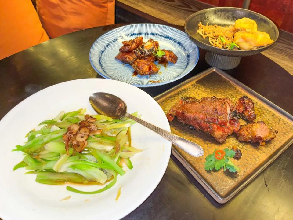 A circular white plate with celery sits next to a square tray of pork. In the background, a blue plate with pieces of breaded cod and a gray tray of fried abalone sits.