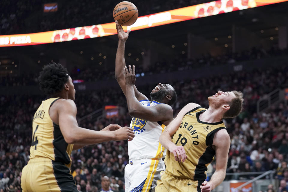 Golden State Warriors forward Draymond Green (23) jumps with the ball while being guarded by Toronto Raptors center Jakob Poeltl, right, and forward Scottie Barnes (4) during the first half of an NBA basketball game Friday, March 1, 2024, in Toronto. (Arlyn McAdorey/The Canadian Press via AP)