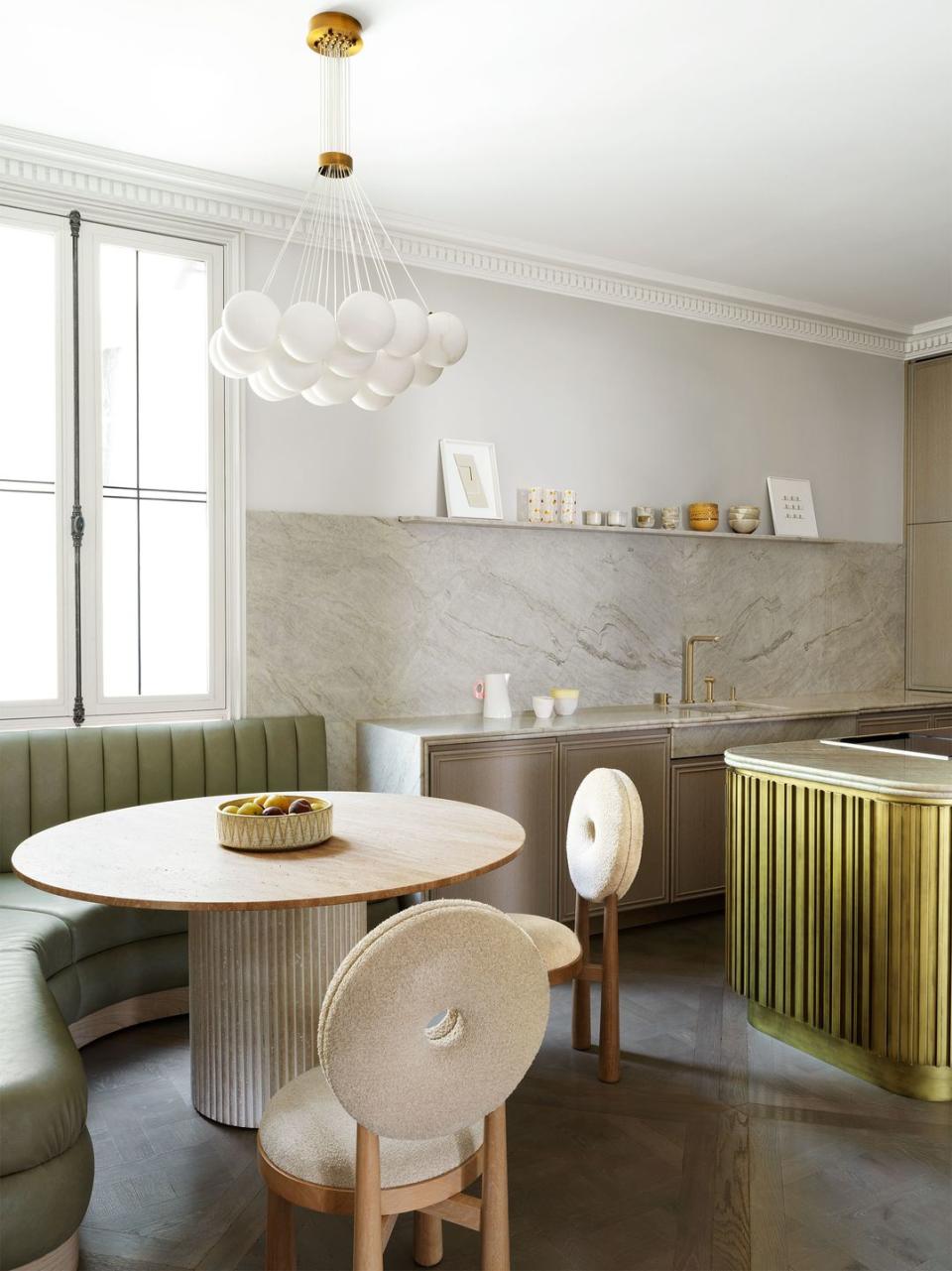 kitchen with beige lower cabinets framed in marble, marble backsplash, open shelf with decorative objects, island with slatted side, pedestal table, wood chairs with circular boucle seats and backs, banquette