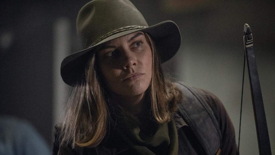 Maggie from The Walking Dead TV show stands in a room wearing a brown hat