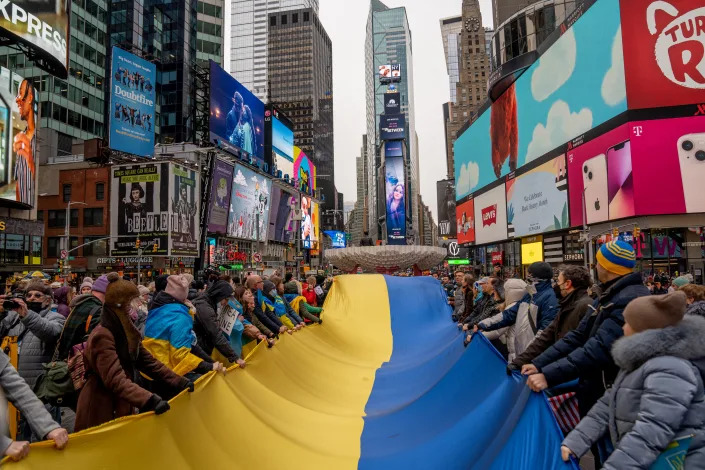 Dozens of protesters line up on each side of a Ukrainian flag many feet long, with theater posters and advertisements of Times Square in the background.