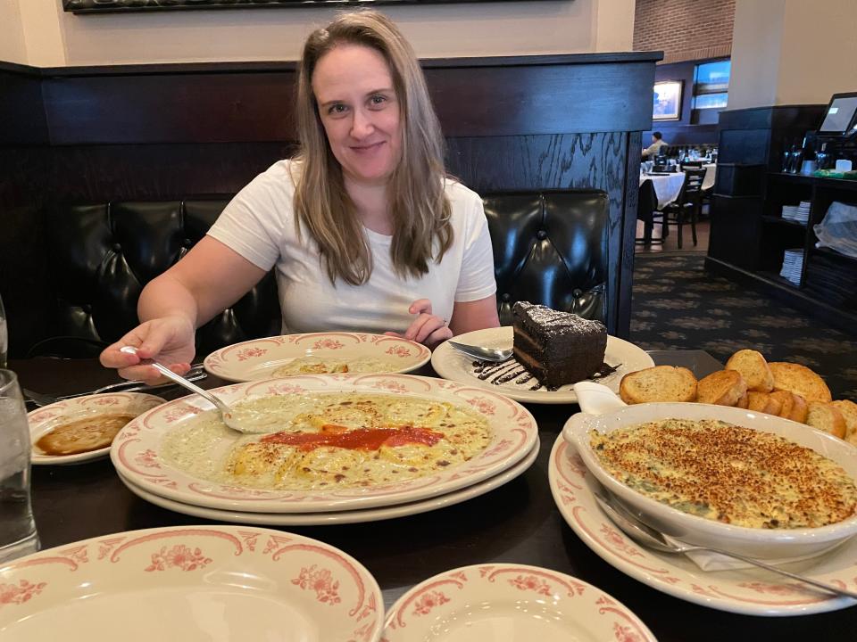 The writer smiles and sits at a table filled with ravioli, a slice of chocolate cake, spinach-artichoke dip, and other plates at Maggiano's