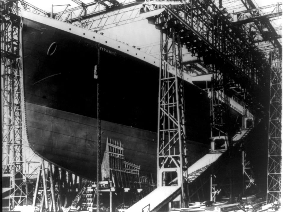 A black-and-white photo showing an exposed part of the Titanic sitting on stocks while poking out of a shipyard.