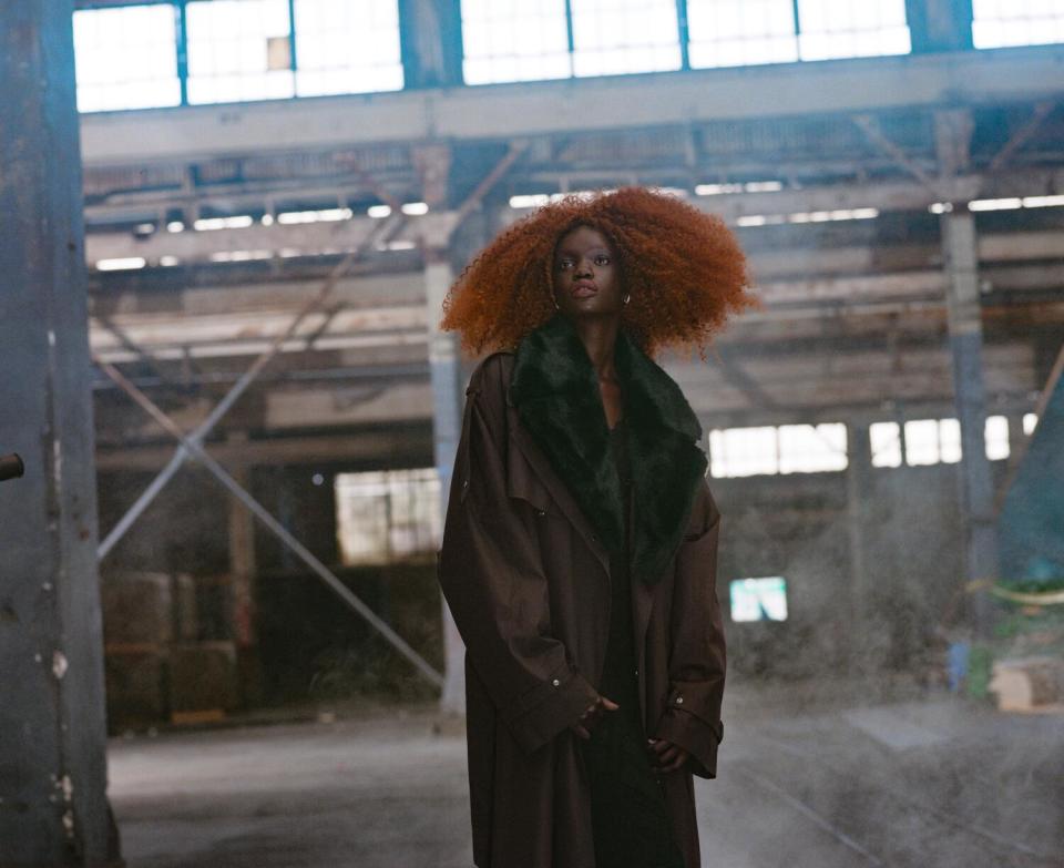 A fashion model stands in an unfinished building, wearing a dark coat