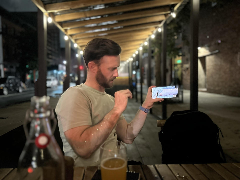 <p>A photo from the iPhone 14's main rear featuring a man holding up a phone with its screen facing the camera and a puzzled expression on his face. The background around him is blurred out.</p>
