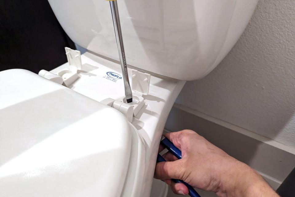 A person unscrewing a toilet seat bolts using a flathead screwdriver and channel lock pliers on a white two-piece ProFlo toilet