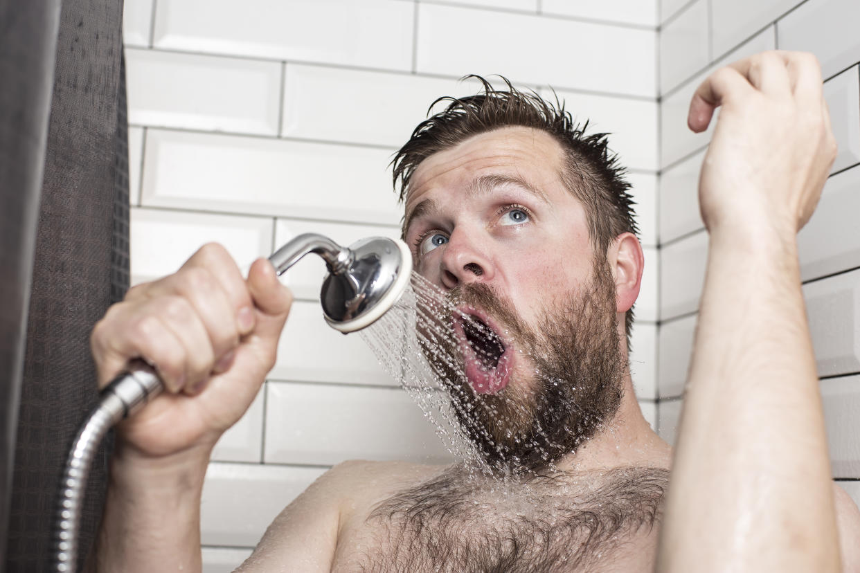 Cute bearded man singing in the bathroom using the shower head with flowing water instead of a microphone. Close-up.