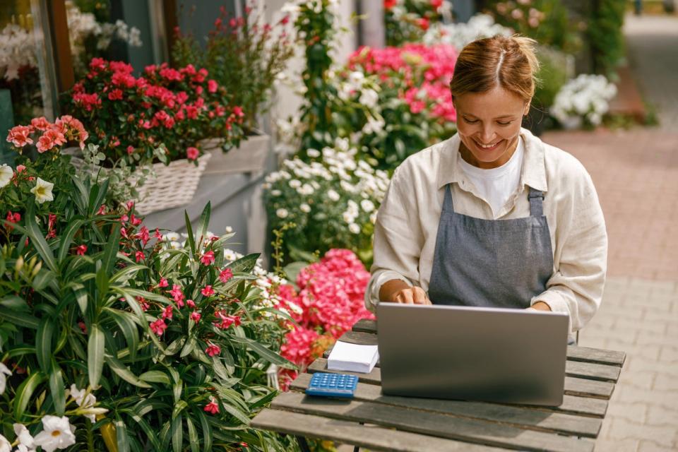 A smiling woman types on a computer, sitting at a table outdoors next to flowers and plants. 