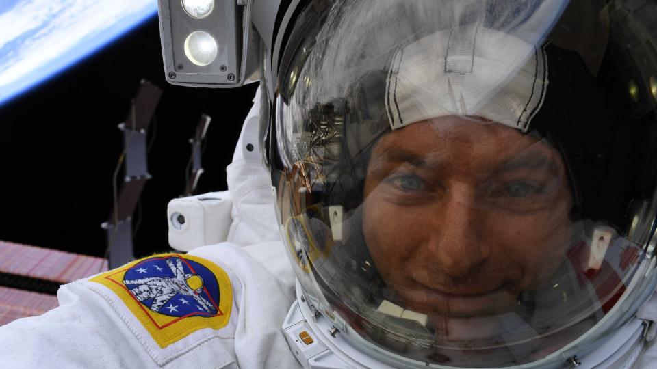 a view of an astronaut in a spacesuit, with helmet visor up. behind are solar panels from the international space station and the black of space