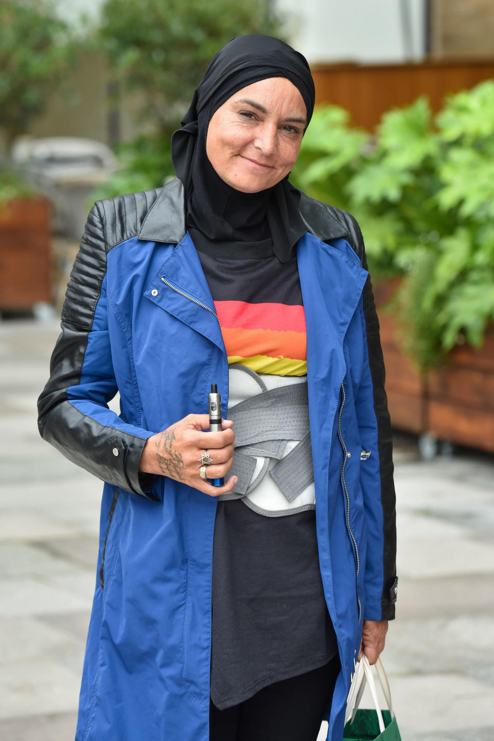 Sinéad O'Connor smiles, wearing a black headscarf, blue trench coat, and black shirt with a graphic design, holding a cigarette and a shopping bag