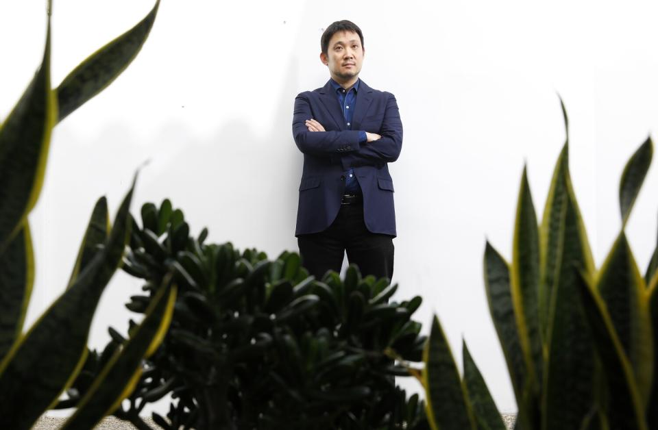 A man stands against a white wall with dark foliage in the foreground of the photo.