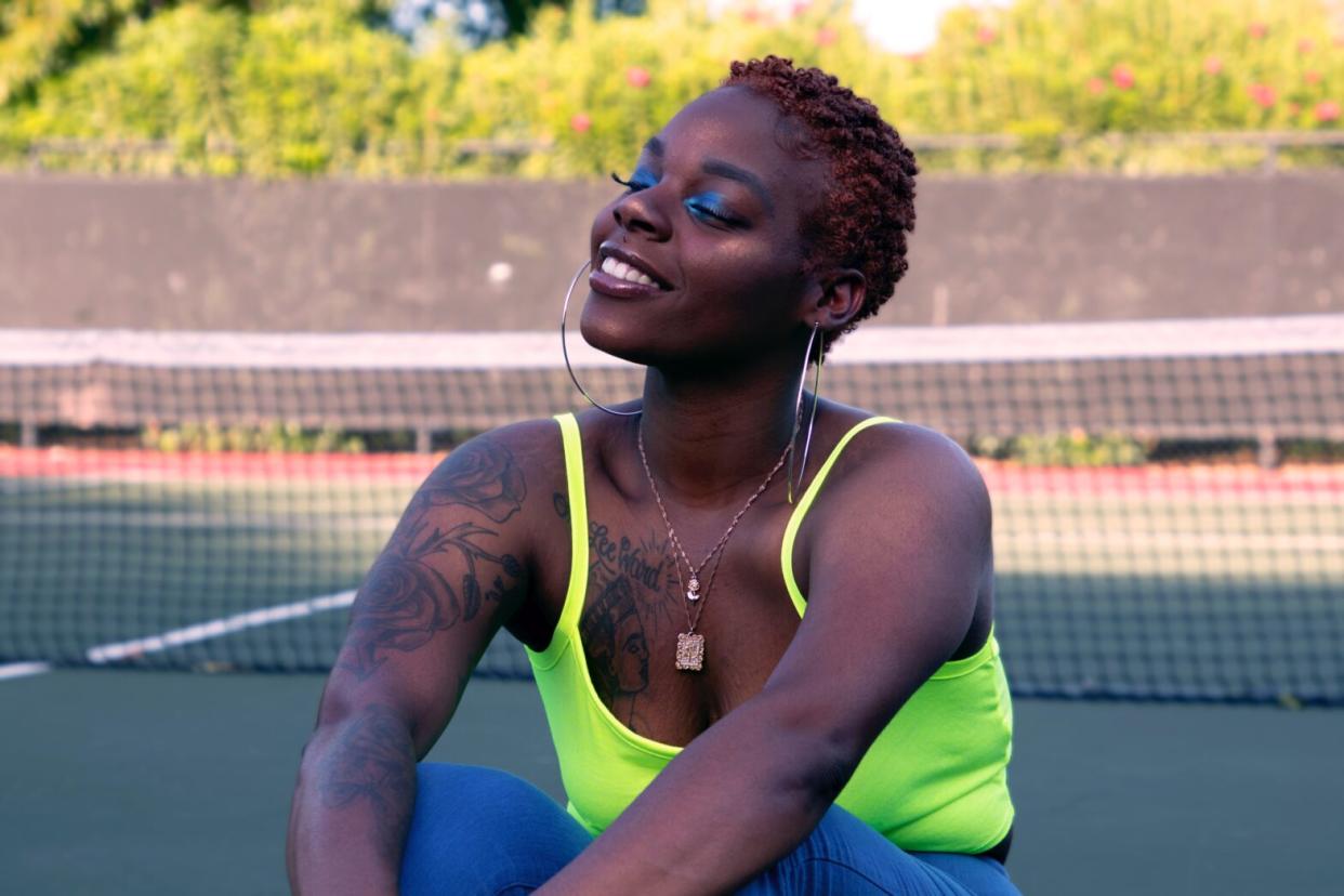 Black woman sitting on a tennis court in a neon green tank top and blue pants smiling wearing silver hoop earrings