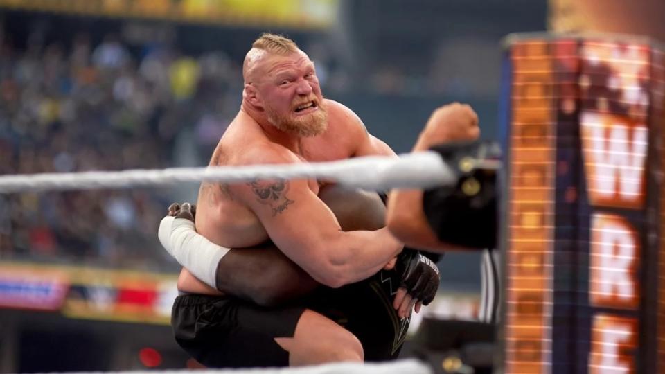 A man with light-toned skin grimaces as he puts a hold on a wrestler with dark-toned skin inside a wrestling ring in front of a large crowd.