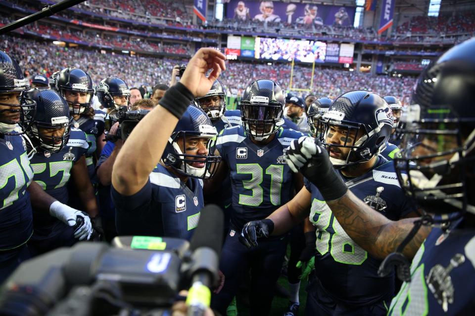 a group of football players gather in a huddle as quarterback russell wilson speaks from the center of them, with his hand raised in the air, all the men wear blue football uniforms and helmets