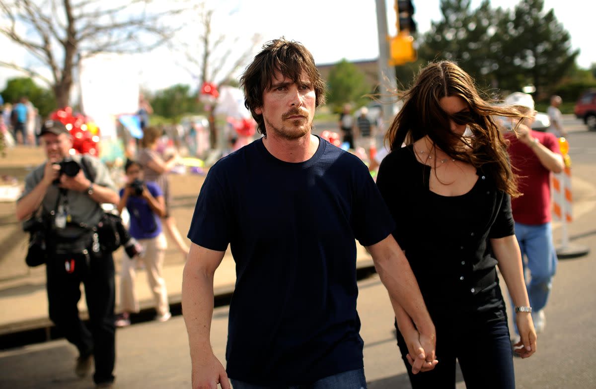 A white man in a blue shirt walks forward outside. Other people are shown behind him. 