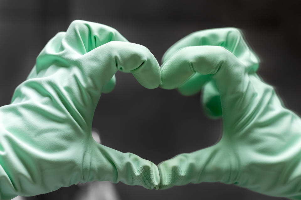 A hand in a mint green rubber glove shows a heart sign with its reflection in the mirror against a gray blurred background. Selective focus. Closeup view