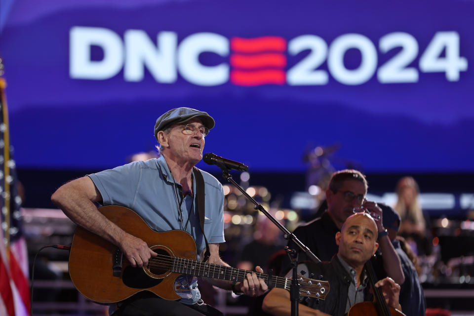 James Taylor tritt mit einer Akustikgitarre auf der Bühne der DNC 2024-Veranstaltung auf. Zwei weitere Musiker spielen im Hintergrund