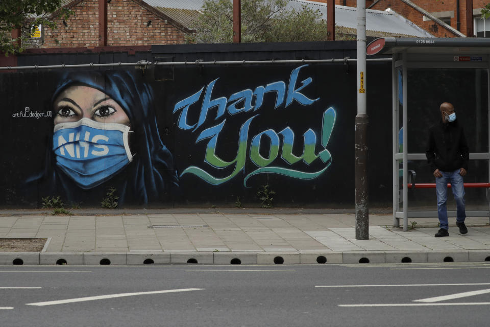 A man wearing a face mask stands at a bus stop next to a recently painted piece of street art by The Artful Dodger (A. Dee) entitled 'NHS Dedication Mural', thanking national health service workers, during the coronavirus lockdown in the Elephant and Castle area of London, Sunday, May 3, 2020. The highly contagious COVID-19 coronavirus has impacted on nations around the globe, many imposing self isolation and exercising social distancing when people move from their homes. (AP Photo/Matt Dunham)