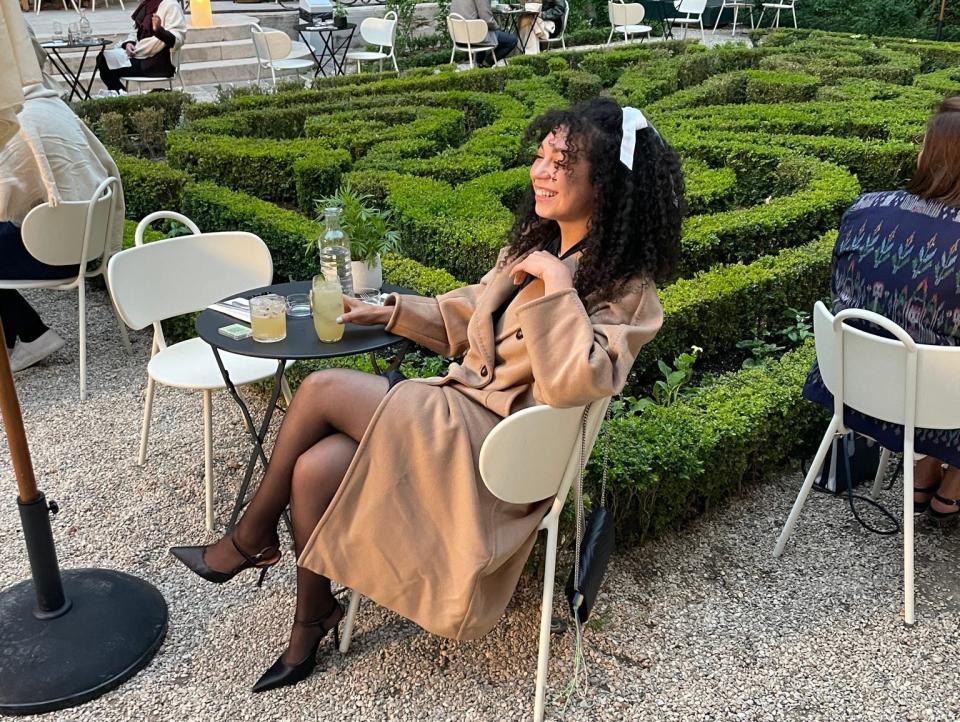 Amber Lucas sits in the gardens of a museum in France wearing a beige trench coat while on a date