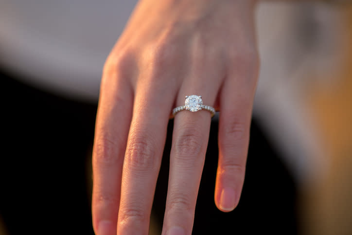 A close-up of a hand wearing a diamond engagement ring with a round-cut gemstone and a band encrusted with smaller diamonds