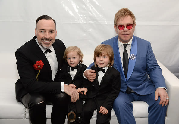 David Furnish, Zachary Jackson Levon Furnish-John, Elijah Joseph Daniel Furnish-John, and Elton John sit on a white couch. David and children wear black suits; Elton wears a blue suit