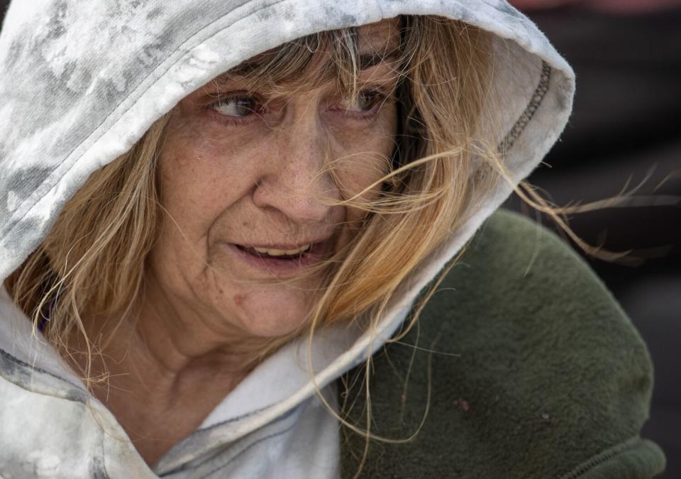 A head-and-shoulders frame of an older woman with blonde hair, wearing a light-grey hooded sweatshirt and a green coat.