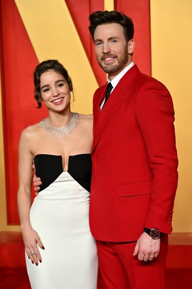 Chris Evans in a red suit and Alba Baptista in a strapless black and white gown, smiling together on the red carpet