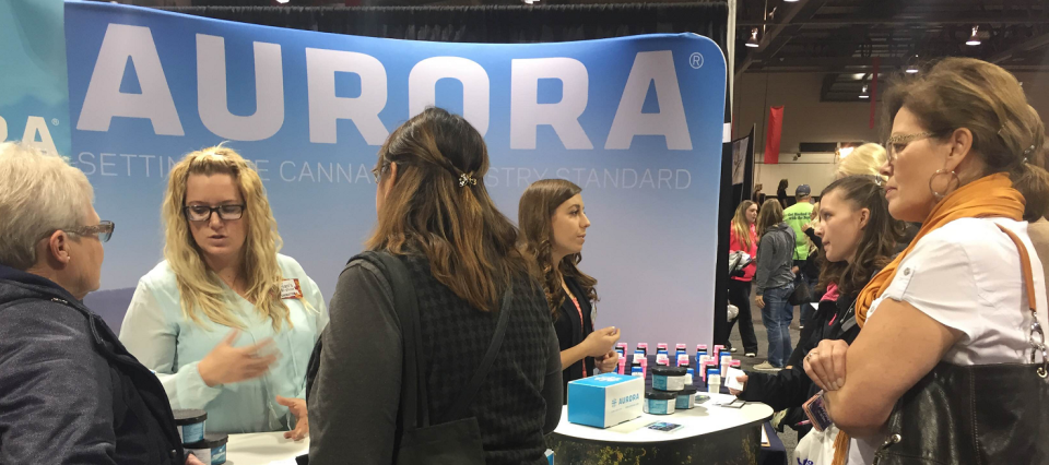 People at a conference booth with the Aurora logo on a sign behind them