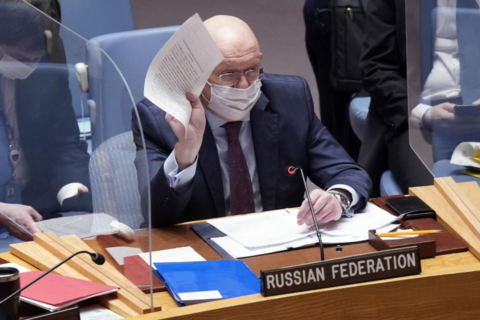 Russia's U.N. Ambassador Vasily Nebenzya sits at a table wearing a face mask and holding up a piece of paper in his right hand.