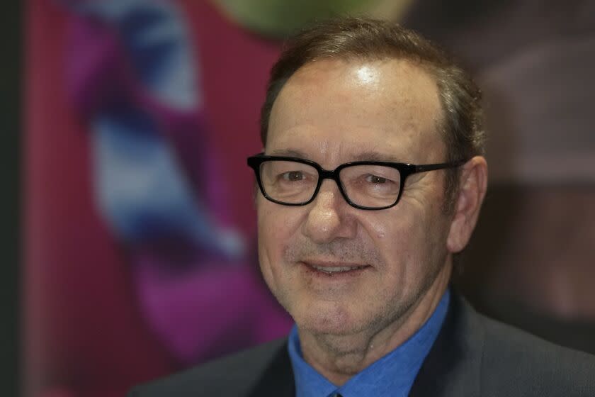 Kevin Spacey wearing thin-framed black glasses, a blue suit shirt and a dark blazer smiling