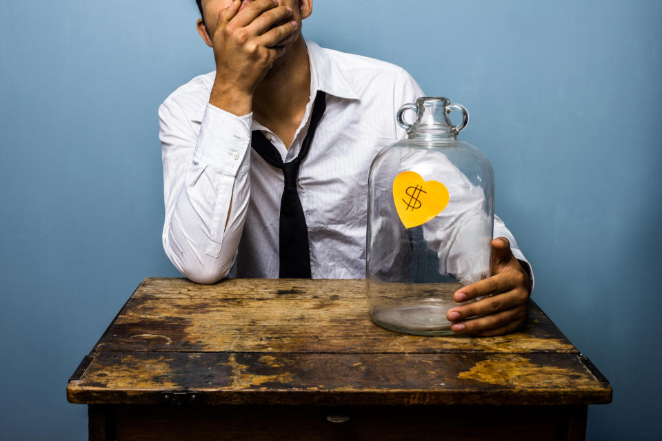 A man is sitting at a small desk with his face in his palm, holding an empty jar with a dollar sign on it.